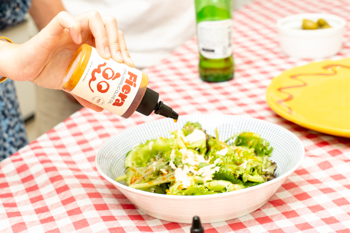 Person pouring a sauce from a bottle labeled 'Fiera' onto a salad on a checkered tablecloth.